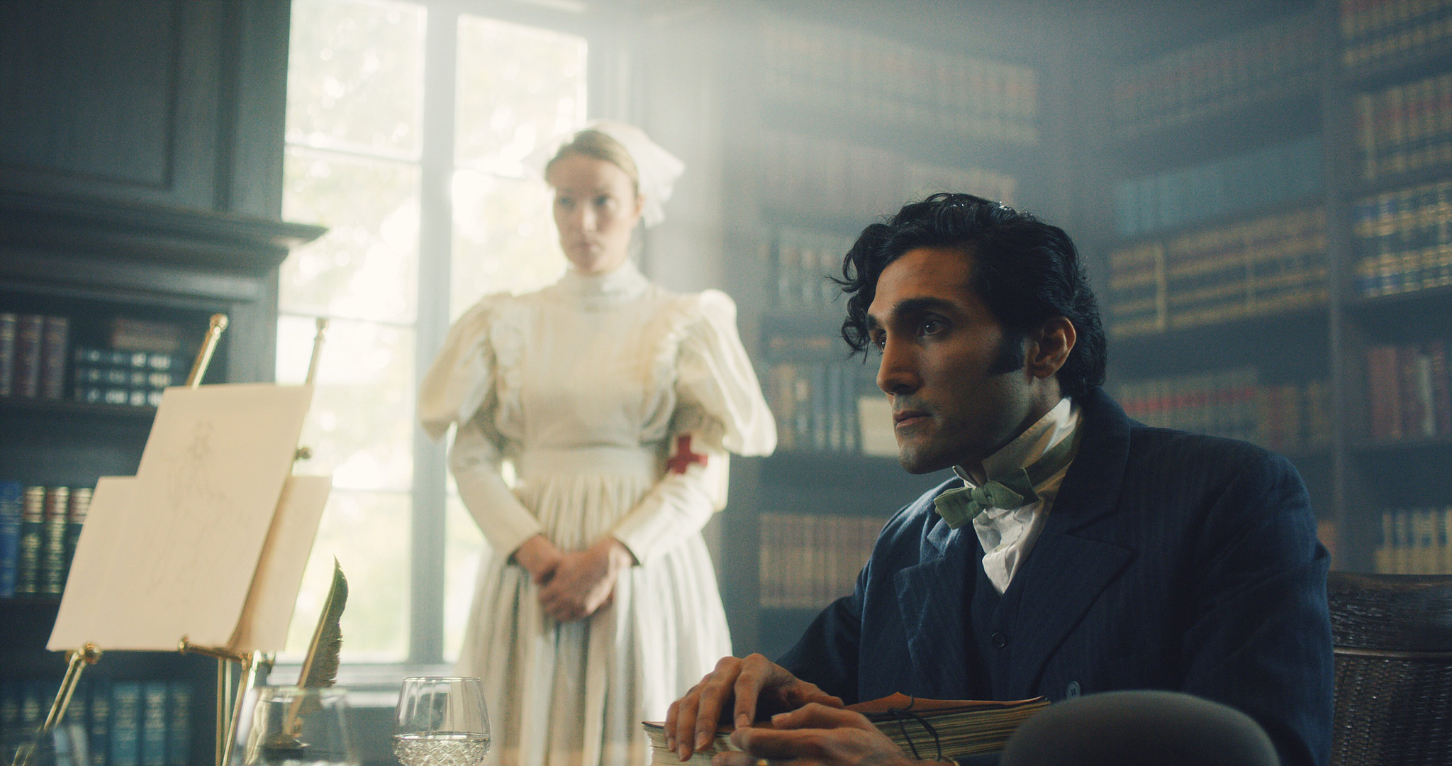 Woman in white at a bright window while a man works at a desk in the asylum library, The Mad Whale