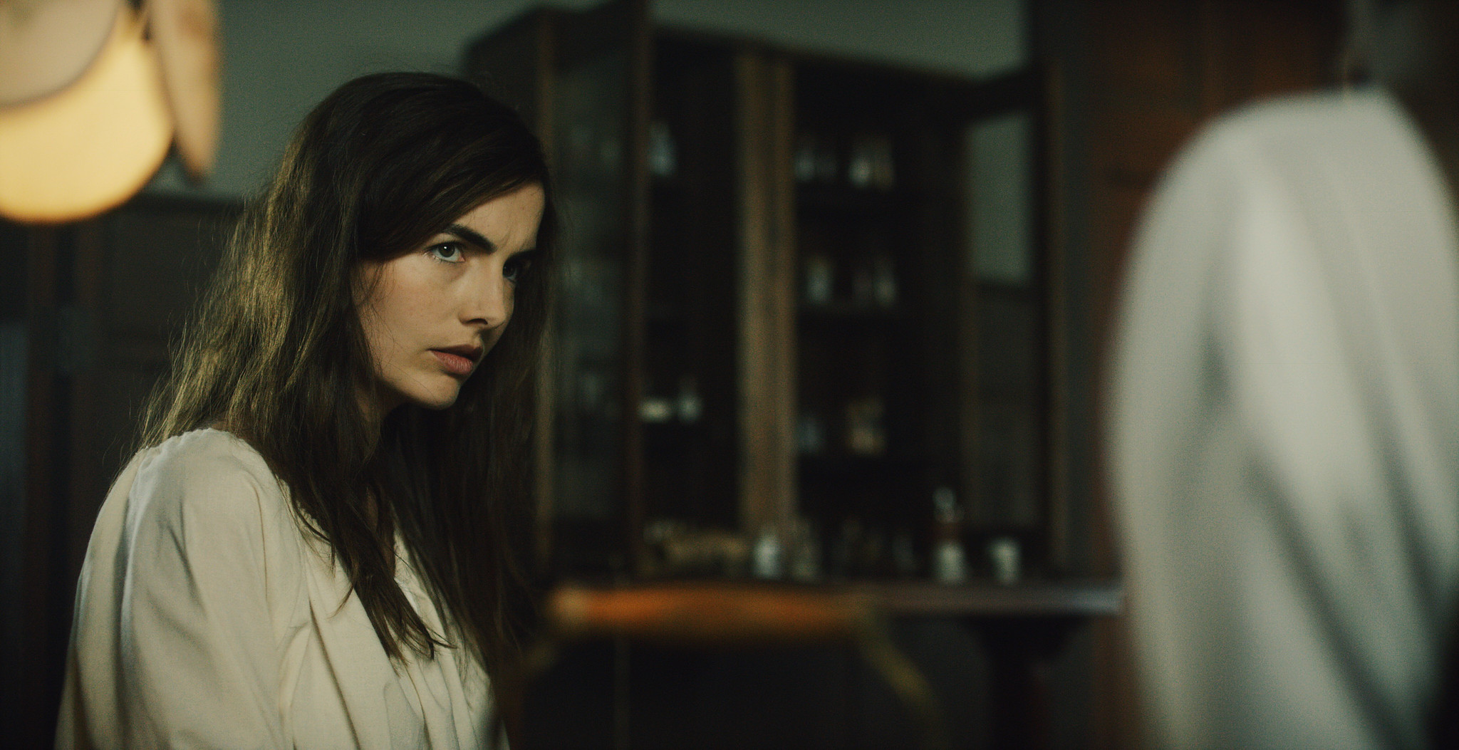 Woman in a pale gown before dark wood cabinetry in a dim asylum room, The Mad Whale
